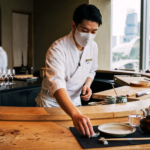 Japanese chef in white uniform and face mask setting up a fine‑dining counter at a luxury restaurant in Singapore with panoramic Marina Bay skyline view.