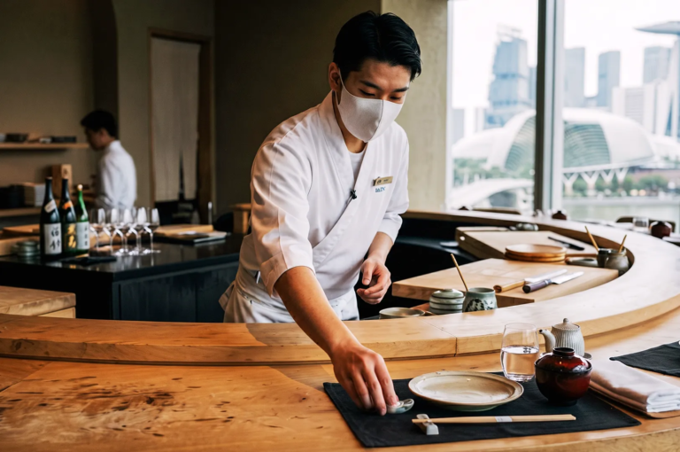 Japanese chef in white uniform and face mask setting up a fine‑dining counter at a luxury restaurant in Singapore with panoramic Marina Bay skyline view.