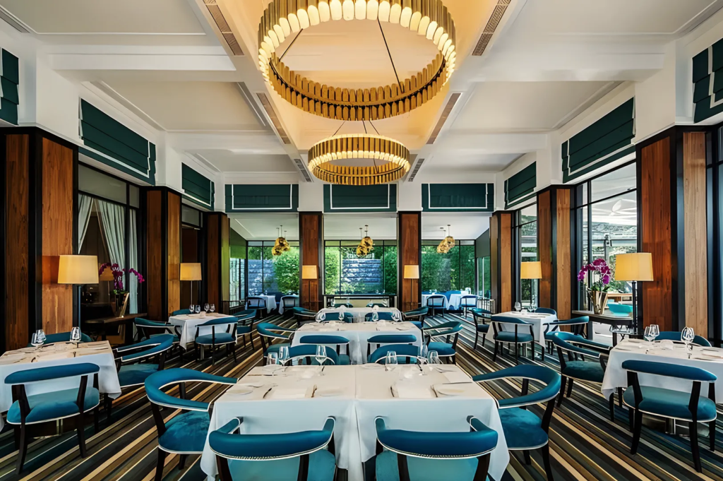 Luxurious restaurant interior with teal‑blue chairs, striped flooring, and golden chandeliers, offering modern fine dining ambiance surrounded by large windows and natural light.