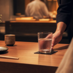 Waiter serving a glass of cold water on a dark coaster at a wooden dining table in a dimly lit Japanese restaurant.