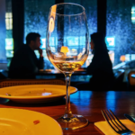 Close‑up of a wine glass with white wine on a restaurant table, with blurred silhouettes of diners and glowing city lights through rain‑streaked windows, capturing cozy nightlife ambiance in a modern urban restaurant.
