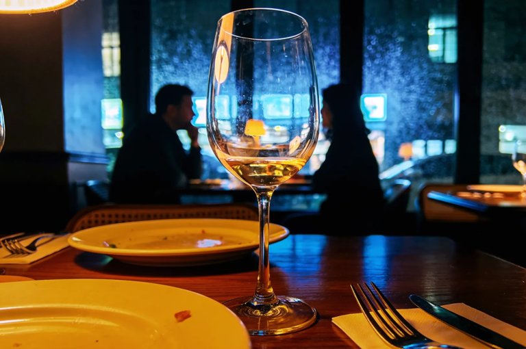 Close‑up of a wine glass with white wine on a restaurant table, with blurred silhouettes of diners and glowing city lights through rain‑streaked windows, capturing cozy nightlife ambiance in a modern urban restaurant.