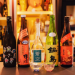 Three clear glasses of sake arranged on a wooden tray with Japanese bottles in the background — traditional sake tasting setup in a Japanese restaurant.