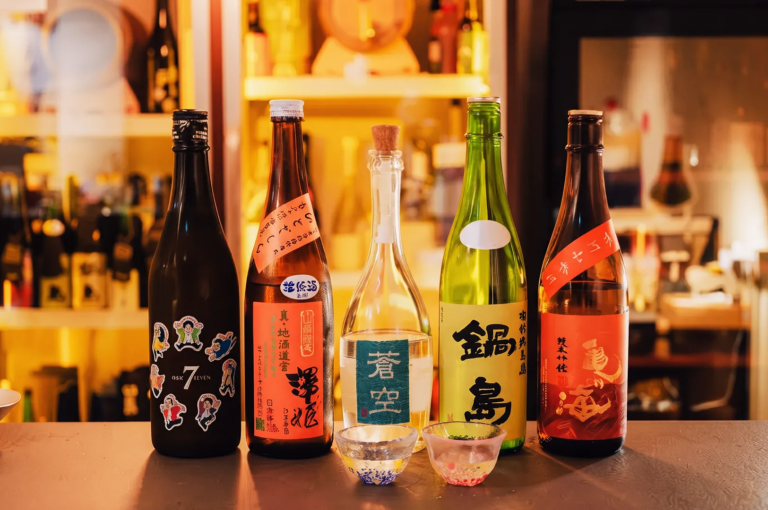 Three clear glasses of sake arranged on a wooden tray with Japanese bottles in the background — traditional sake tasting setup in a Japanese restaurant.