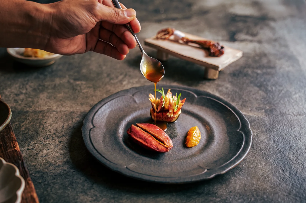 Chef drizzling sauce over seared meat and vegetables on a dark ceramic plate, capturing the precision and artistry of fine dining plating.