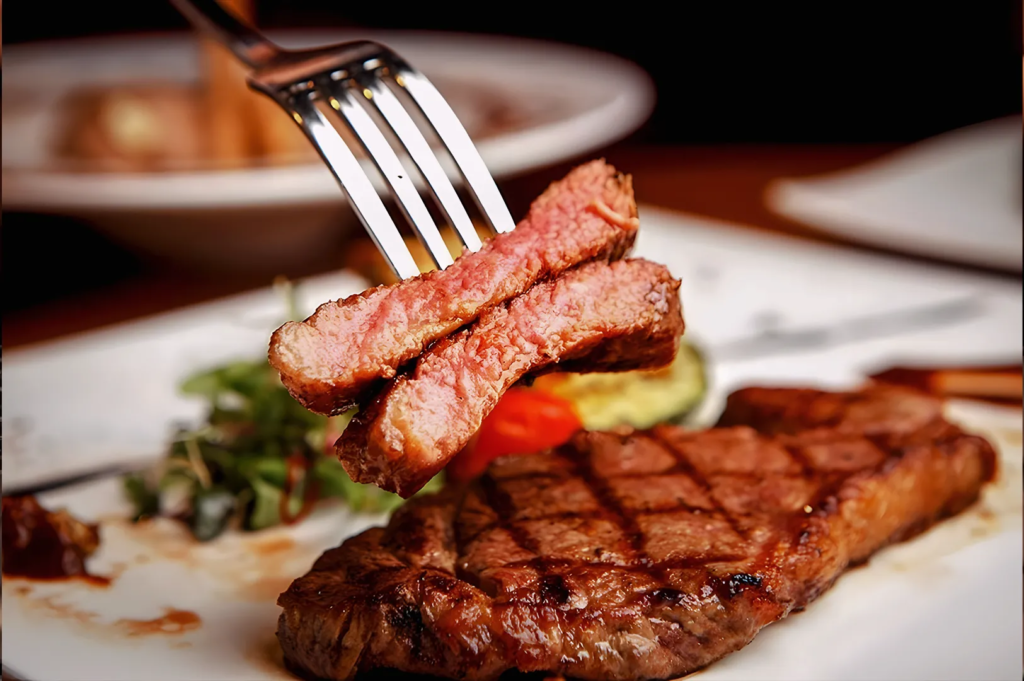 Close‑up of tender steak slices held by a fork, showing juicy pink interior and charred grill marks; fine‑dining steak detail.