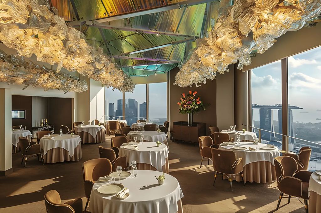 Fine dining restaurant with panoramic view of Marina Bay Sands and ArtScience Museum, featuring elegant round tables and sculptural ceiling lights.
