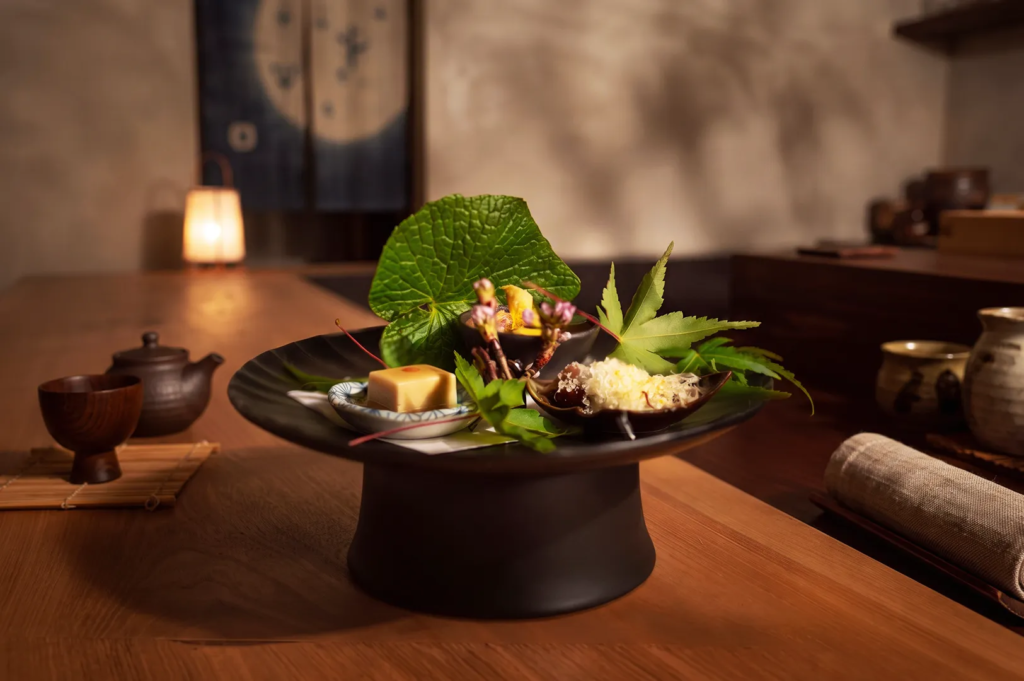 Traditional kaiseki dish with tofu, vegetables, and fresh leaves served on a black pedestal plate in a Japanese restaurant.