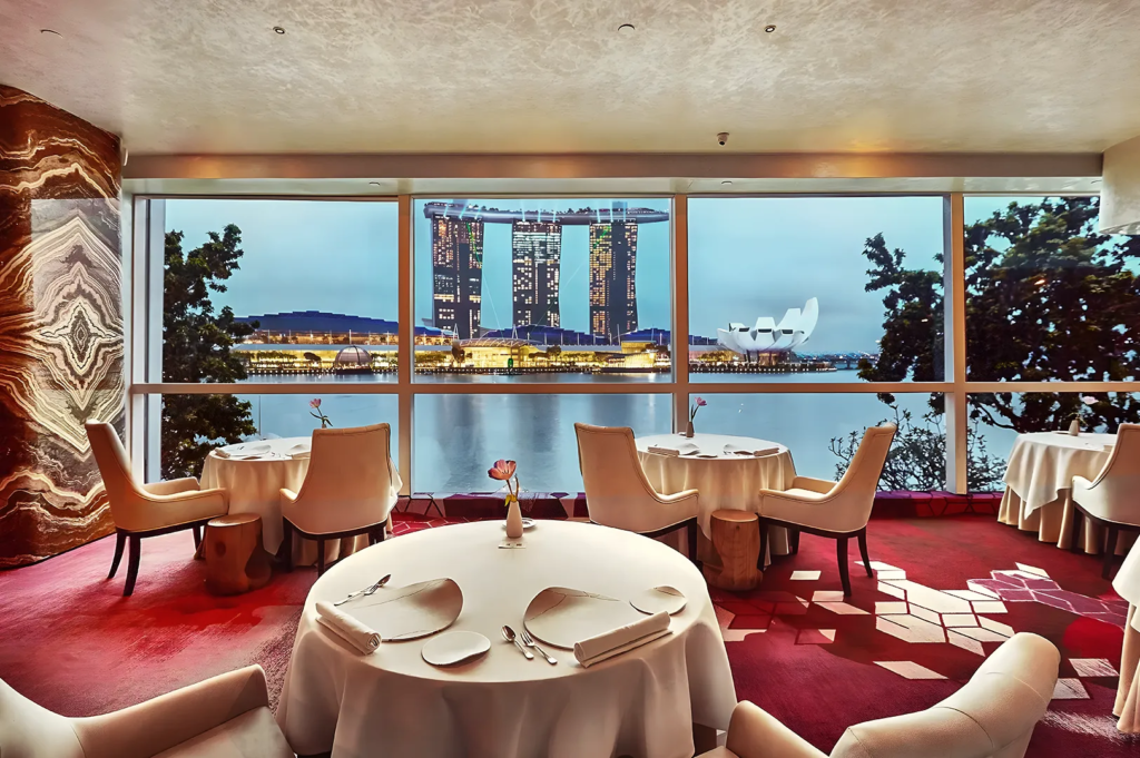 Restaurant overlooking Marina Bay with white‑clothed tables and beige chairs, offering scenic views of Singapore’s skyline and Marina Bay Sands.
