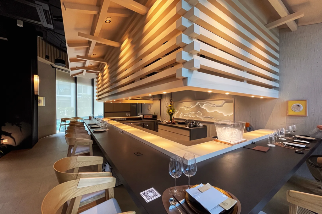 Contemporary omakase dining space with geometric ceiling beams and open kitchen design in Singapore.