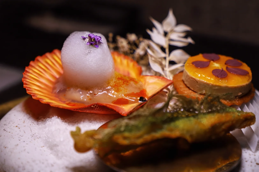 Artistic Japanese dish with scallop foam, edible flowers, and layered mousse presented in a shell at a Michelin‑style restaurant.