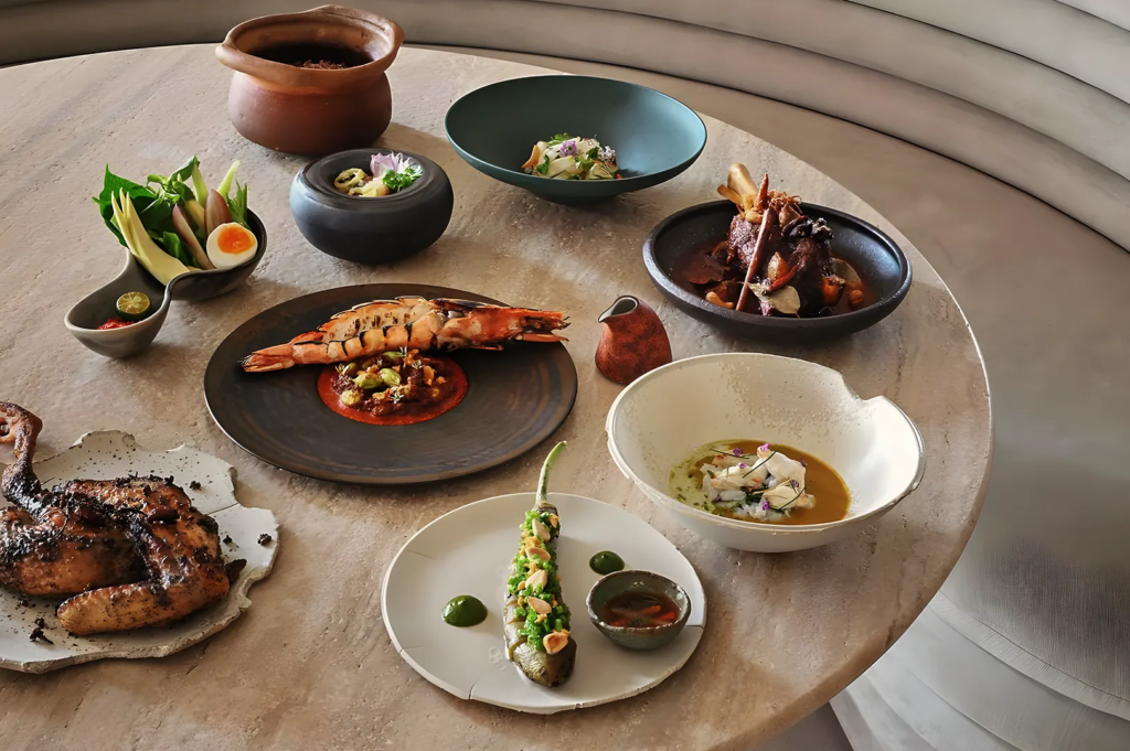 Assorted fine dining dishes including grilled prawn, roasted chicken, stuffed chili, and vegetable sides, showcasing modern Asian fusion cuisine.