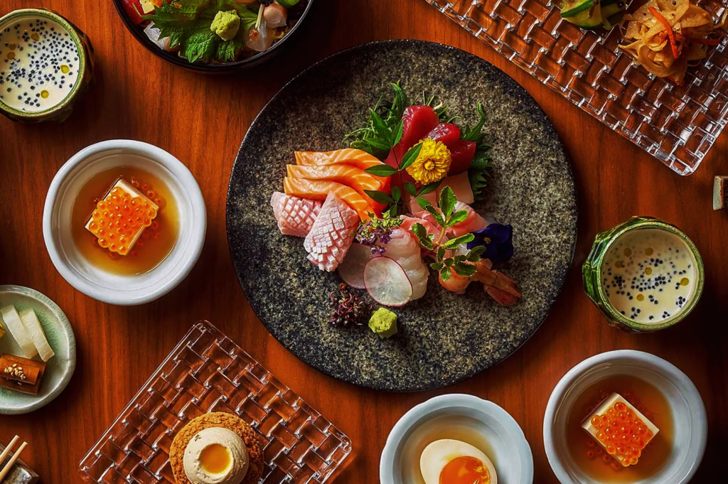 Colorful sashimi platter with salmon, tuna, and tofu topped with ikura surrounded by Japanese side dishes on a wooden table.