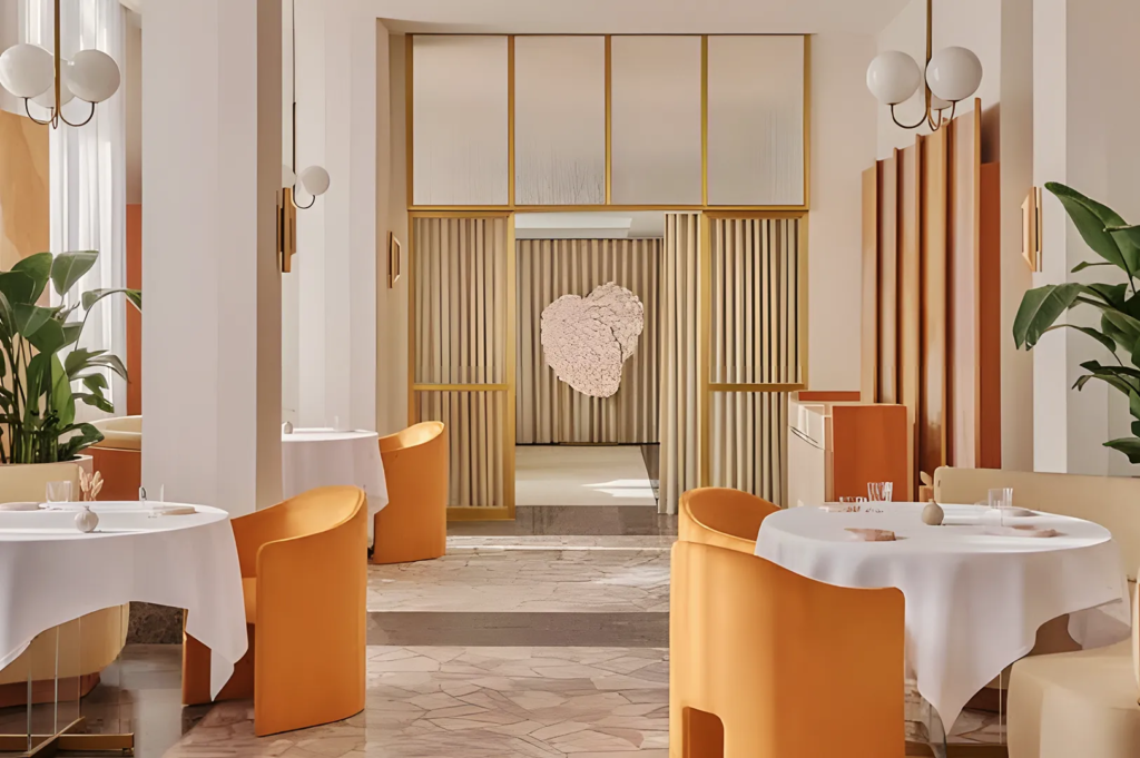 Modern fine dining restaurant with warm beige and orange tones, marble flooring, and sculptural wall art, featuring elegant round tables and gold accents.