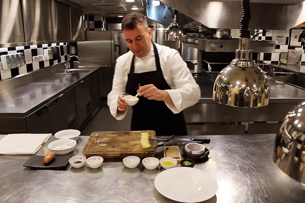 Chef in white uniform and black apron preparing gourmet ingredients in a modern stainless‑steel kitchen at Les Amis Singapore, highlighting precision and culinary artistry.