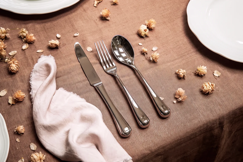 Close‑up of polished silver cutlery on brown linen tablecloth with dried floral accents — refined dining table arrangement for luxury restaurant.