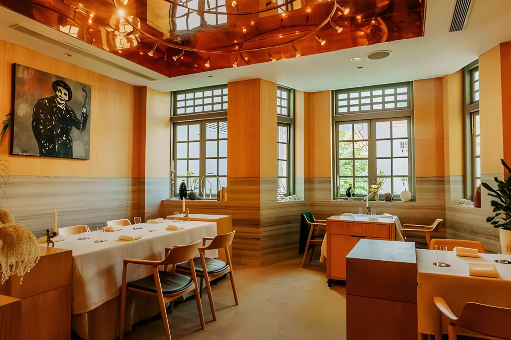Elegant fine‑dining restaurant with large grid windows, copper ceiling accents, and minimalist wooden furniture. Tables are set with white linens and refined tableware, exuding a luxurious yet artistic ambiance perfect for contemporary gastronomy.