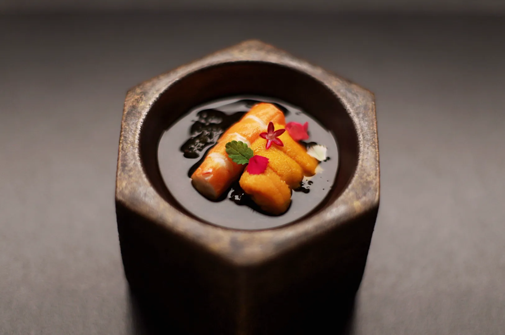 Gourmet uni and shrimp dish served in a hexagonal bowl with dark sauce and edible flowers at a Michelin‑style Japanese restaurant.