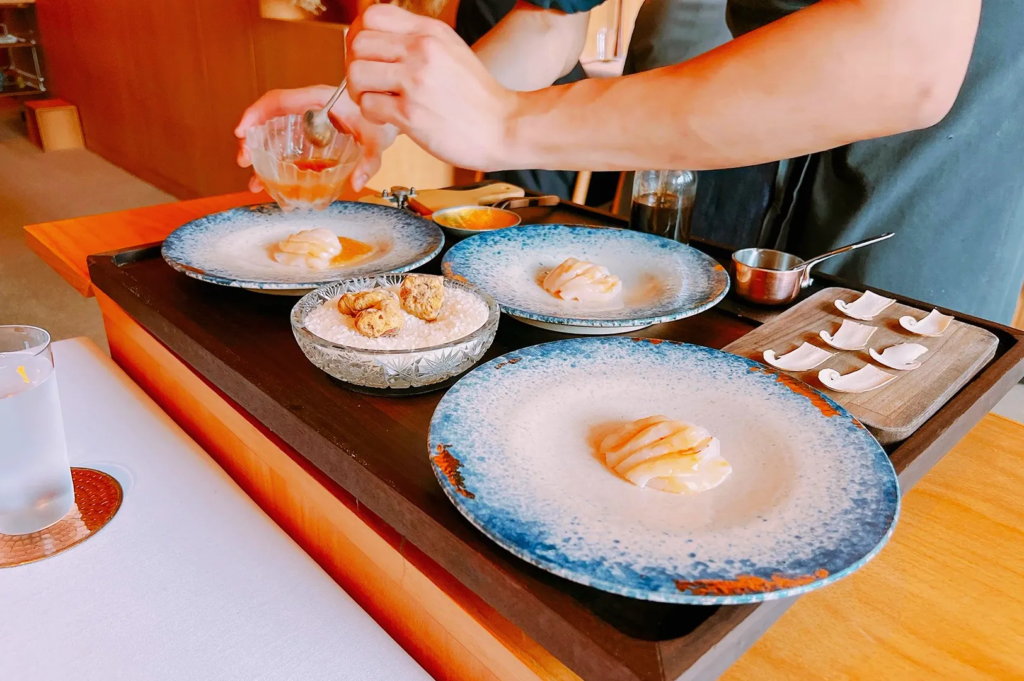 Chef plating gourmet seafood dishes on blue‑rimmed ceramic plates in a high‑end restaurant. The scene highlights meticulous culinary presentation with fresh ingredients, sauces, and truffle garnishes, showcasing modern Japanese‑inspired fine dining.