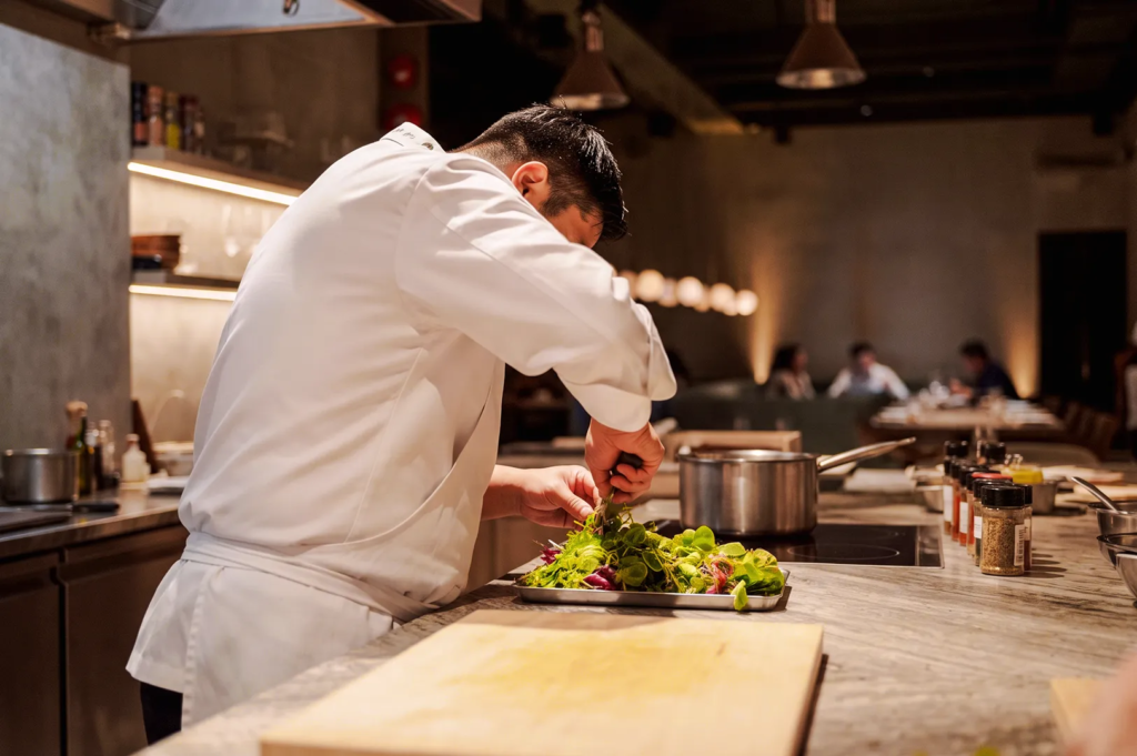 Professional chef in a modern restaurant kitchen carefully plating fresh green salad with herbs on a marble countertop, surrounded by spices and cookware under warm ambient lighting.