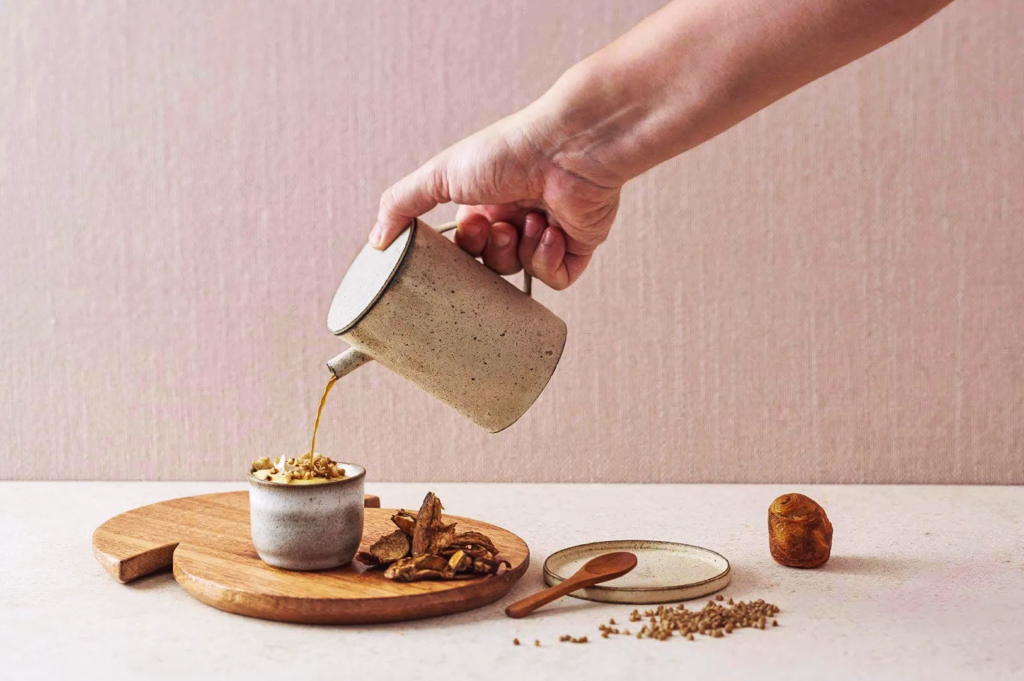 Hand pouring broth into ceramic cup with dried mushrooms and rustic tableware — refined culinary presentation at Odette restaurant Singapore.