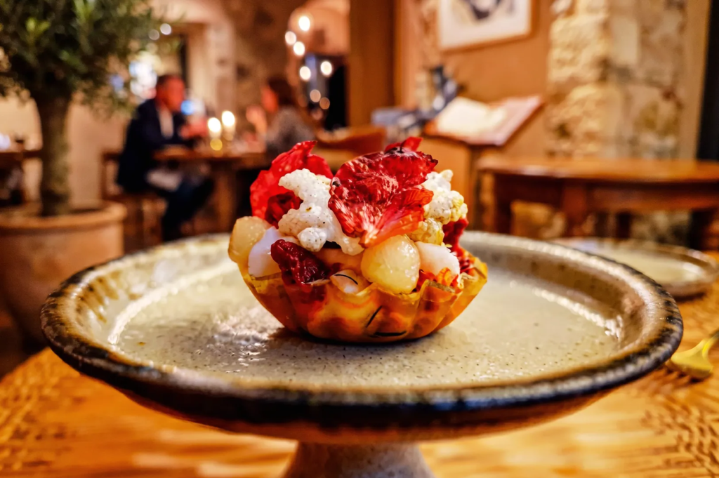 Close‑up of a colorful gourmet dessert served in an edible shell bowl, featuring vibrant red, white, and yellow elements like dehydrated fruits and meringue crisps. The rustic restaurant background adds warmth and artisanal charm to the fine‑dining experience.
