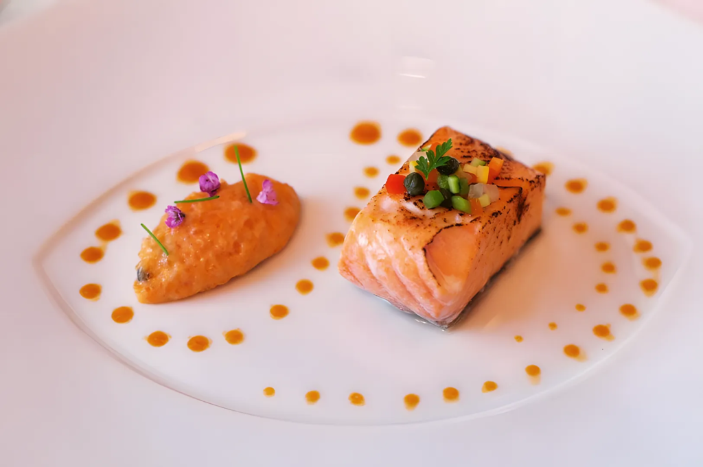 Gourmet seared salmon topped with diced vegetables and capers, plated with orange puree and decorative sauce dots on a white fine dining plate.