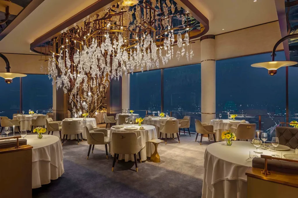 Luxurious Michelin‑star restaurant interior featuring round tables, white linens, and a stunning crystal chandelier overlooking a night cityscape.