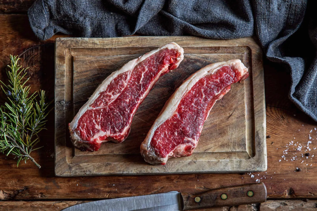 Two marbled new york strip steaks on a wooden board with rosemary and coarse salt; premium beef prepared for cooking.