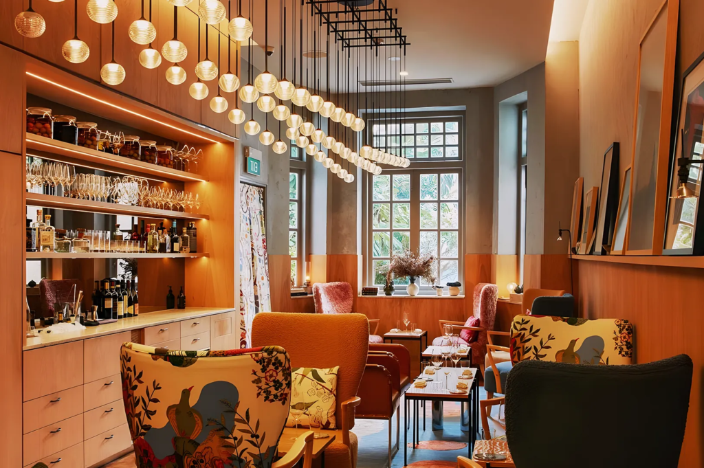 Stylish café or bar interior with artistic armchairs, pendant globe lights, and shelves filled with wine and spirits. The mix of floral upholstery, soft lighting, and modern décor creates a chic, inviting space for social gatherings or afternoon drinks.