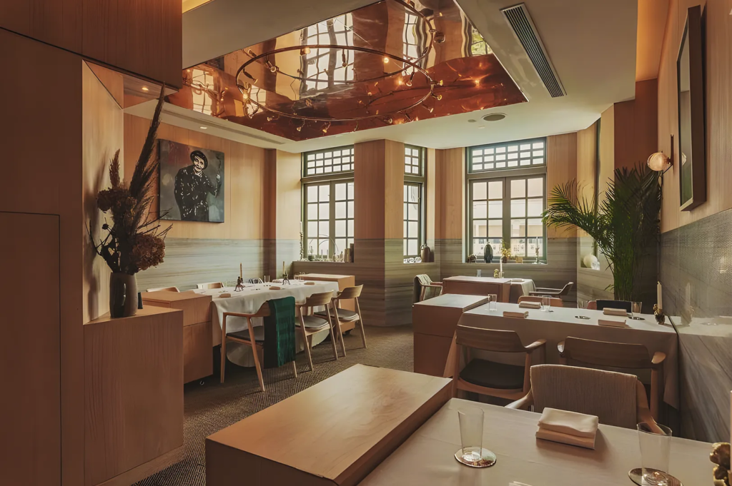 Contemporary restaurant with copper ceiling panels, wooden furniture, and large windows, blending natural light with elegant minimalist design.