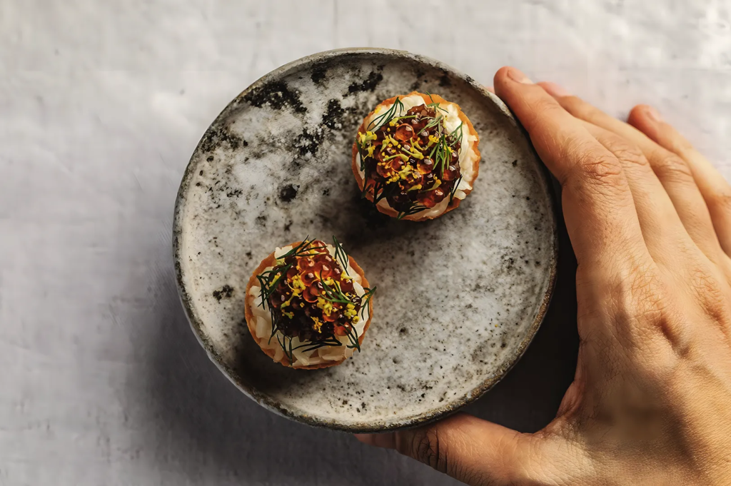 Close‑up of two bite‑sized gourmet appetizers topped with salmon roe, herbs, and edible flowers on a rustic ceramic plate, highlighting fine dining artistry.