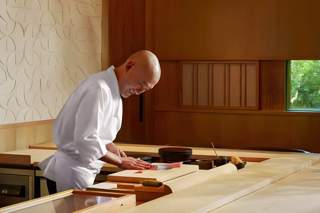 Japanese chef smiling while preparing sushi behind a wooden counter in a serene fine‑dining restaurant.