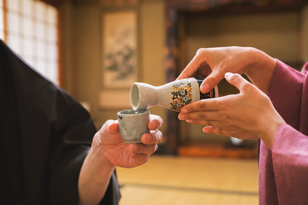 Close‑up of hands pouring sake from a floral ceramic flask into a cup during a Japanese tea‑room ceremony — authentic cultural sake serving tradition.