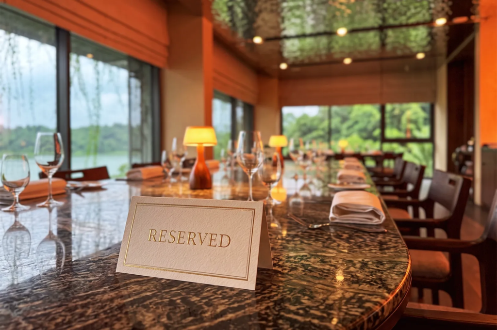 Gold embossed reserved card on a polished dark marble dining table, elegantly set with wine glasses, warm table lamps, and scenic window views of lush greenery.