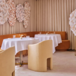 Sophisticated restaurant interior with beige and caramel tones, round tables draped in white linens, and artistic wall installations, showcasing modern luxury dining ambiance.