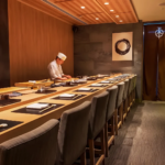 Elegant omakase sushi restaurant interior with a Japanese chef preparing dishes behind a minimalist wooden counter, featuring warm lighting and refined décor for an authentic dining experience.