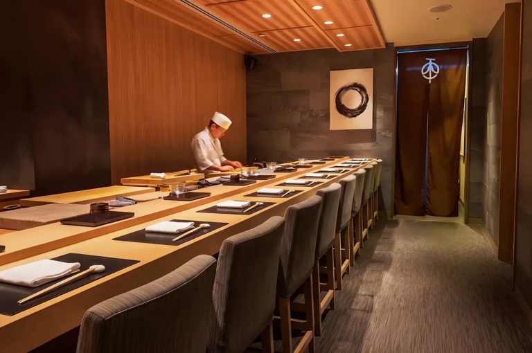 Elegant omakase sushi restaurant interior with a Japanese chef preparing dishes behind a minimalist wooden counter, featuring warm lighting and refined décor for an authentic dining experience.