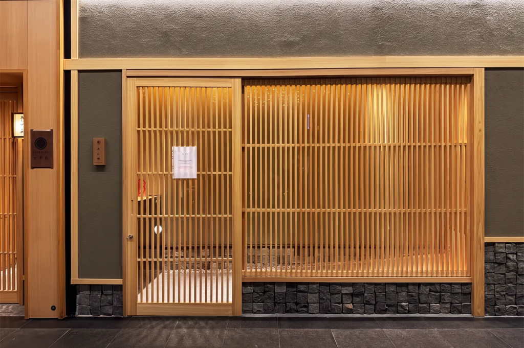 Elegant Japanese restaurant exterior in Singapore featuring wooden lattice doors, stone base accents, and warm ambient lighting that highlights minimalist architecture.