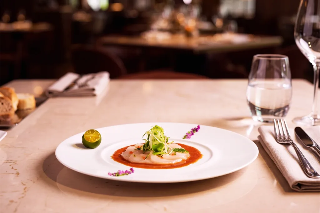 Elegant fine‑dining dish served at a luxury restaurant in Singapore featuring scallop carpaccio with tomato sauce, microgreens, edible flowers, and lime garnish on a marble table.