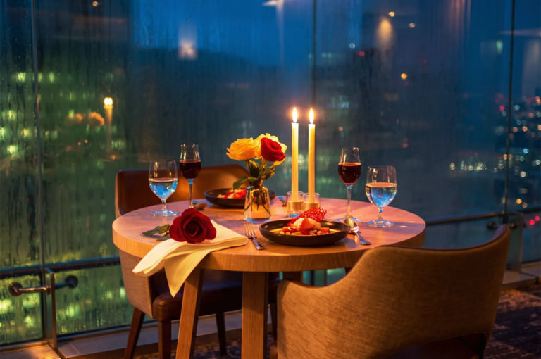 Elegant table setup for two featuring wine glasses, plated desserts, roses, and glowing candles against a misty glass window overlooking Singapore’s night skyline, perfect for a romantic fine‑dining experience.