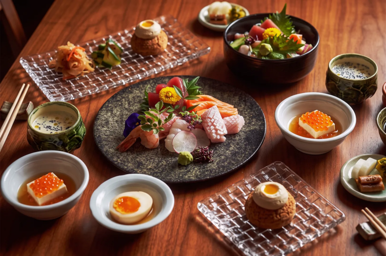 Elegant assortment of fresh sashimi including salmon, tuna, squid, and shrimp, beautifully plated with edible flowers, radish slices, and wasabi on a stone plate surrounded by side dishes and tea cups.