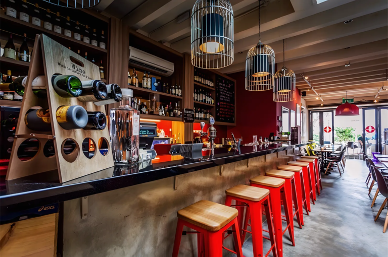 Modern wine bar interior in Singapore featuring red stools, black countertop, hanging cage lights, and wooden wine rack with bottles displayed stylishly.