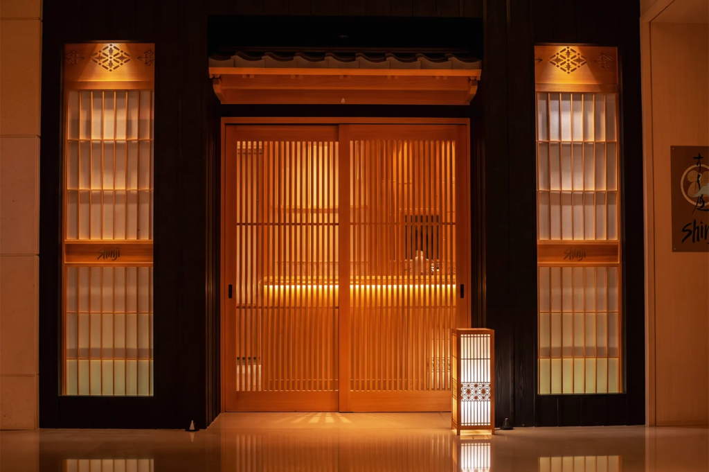 Elegant entrance of Shinji Japanese restaurant in Singapore featuring traditional wooden sliding doors, warm lighting, and minimalist Japanese architecture.