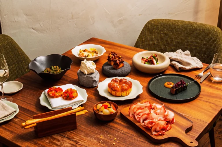 Elegant wooden table set with assorted small plates, charcuterie, breadsticks, and artistic dishes, showcasing modern European cuisine and refined plating in a cozy dining setting.
