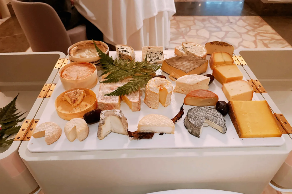 Curated assortment of gourmet cheeses including soft, aged, and blue varieties displayed on a white tray with decorative ferns, representing premium European cheese tasting experience.