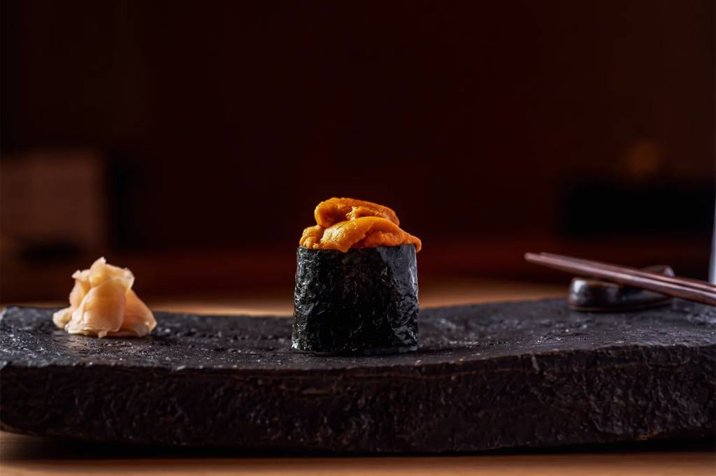 Close‑up of uni sushi served on a black stone plate with pickled ginger and chopsticks, showcasing premium Japanese sea urchin delicacy in fine‑dining presentation.