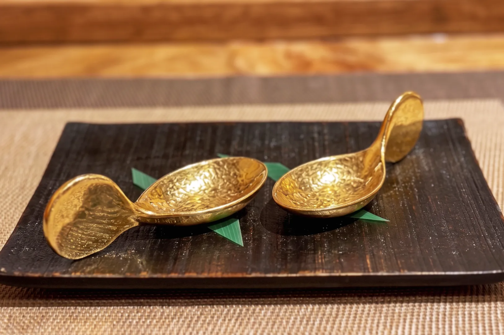 Two empty decorative gold tasting spoons on a dark wooden platter at an omakase dining counter.