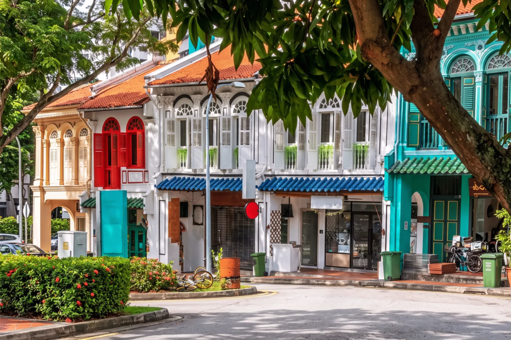 Colorful heritage shophouses in Singapore with vibrant facades, arched windows, and red, blue, and green shutters surrounded by tropical greenery.