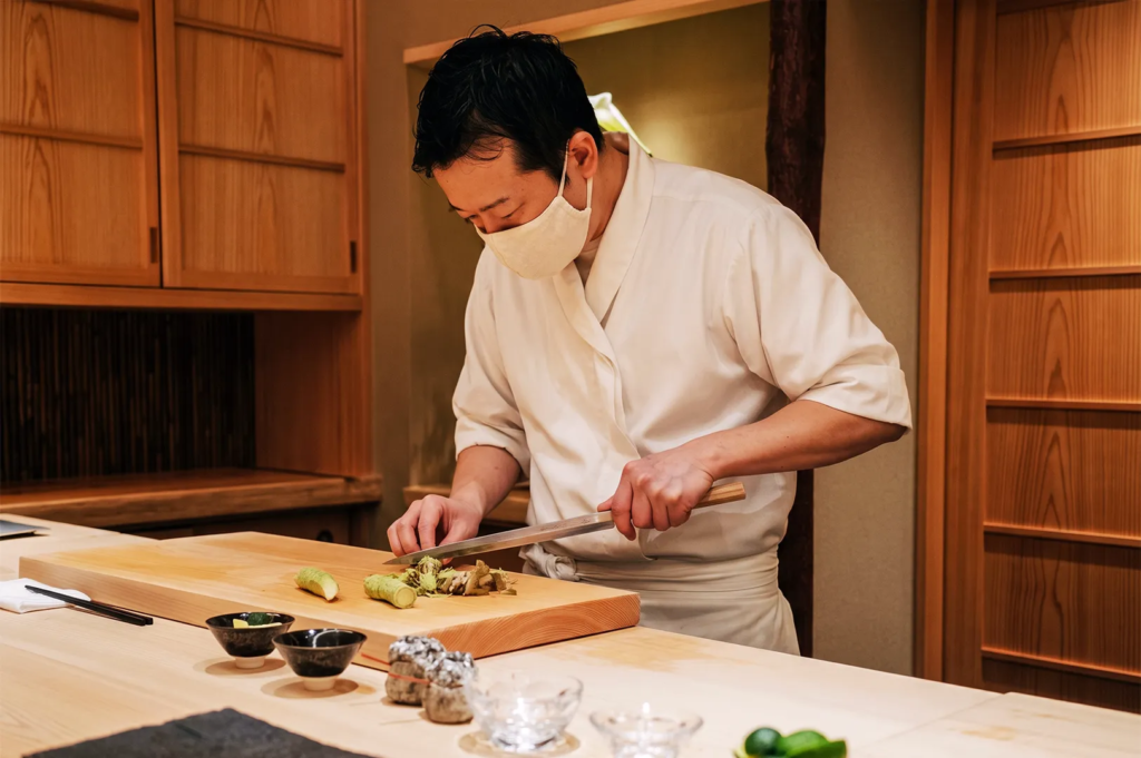 Japanese sushi chef in white uniform preparing fresh wasabi at a wooden counter inside a traditional omakase restaurant in Singapore.